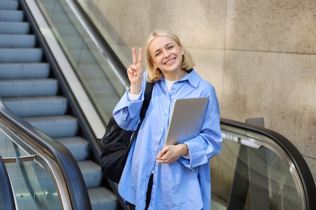 Photo gratuite une belle étudiante souriante avec un ordinateur portable et un sac à dos utilisant l'escalator dans la ville montrant la paix