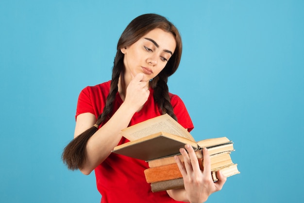 Belle étudiante en t-shirt rouge lisant attentivement un livre sur un mur bleu.