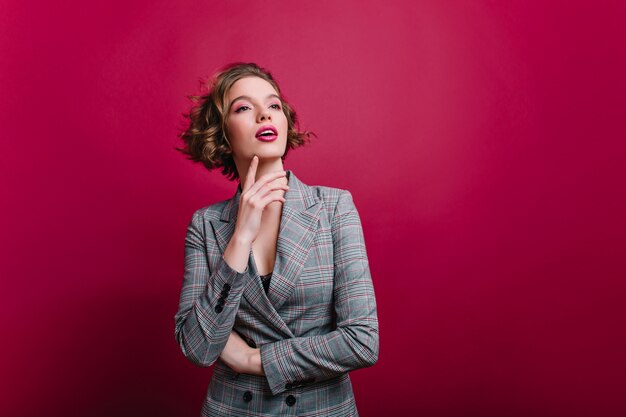 Belle femme d'affaires confiante posant sur un mur bordeaux et regardant ailleurs. Rêveuse fille caucasienne avec coupe courte en veste grise debout