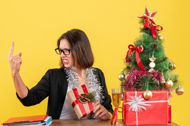 Belle femme d'affaires en costume avec des lunettes pointant vers le haut avec colère et assis à une table avec un arbre de Noël dessus dans le bureau sur jaune