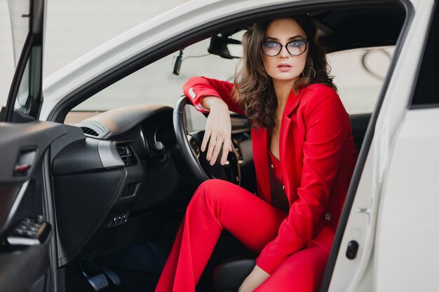 Belle femme d'affaires riche sexy en costume rouge assis dans une voiture blanche, portant des lunettes