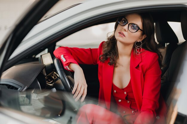 Belle femme d'affaires riche sexy en costume rouge assis dans une voiture blanche, portant des lunettes