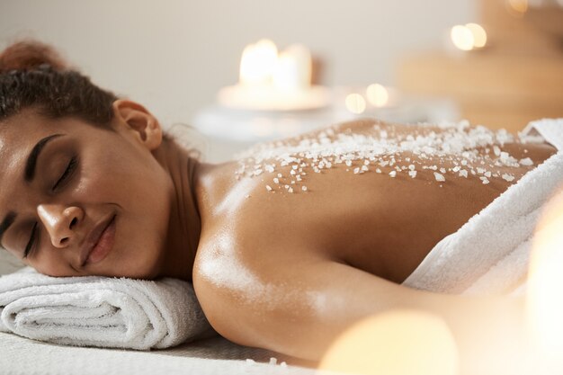 Belle femme africaine au repos relaxante avec du sel de mer sur le dos dans le salon spa.