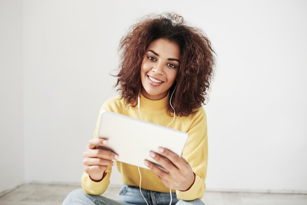 Belle femme africaine dans les écouteurs souriant tenant la tablette assis sur le sol