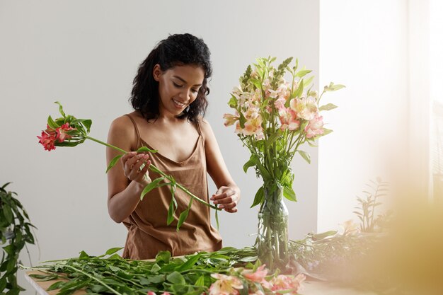 Belle femme africaine souriante faisant un bouquet d'alstroemerias au lieu de travail sur mur blanc.