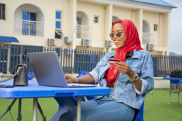 Belle femme africaine souriante tout en utilisant son ordinateur portable et sa carte de crédit en plein air.