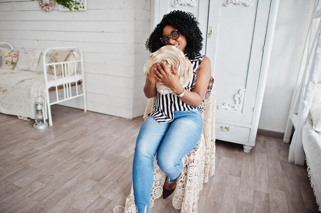 Photo gratuite belle femme afro-américaine avec des cheveux afro bouclés et des lunettes posées dans la chambre assise sur une chaise rétro avec une rose décorative dans les mains