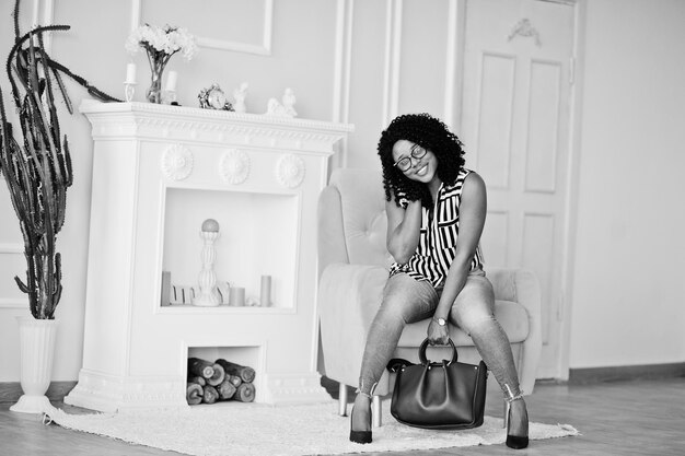 Belle femme afro-américaine avec des cheveux afro bouclés et des lunettes sac à main posé dans la chambre assis sur une chaise jaune