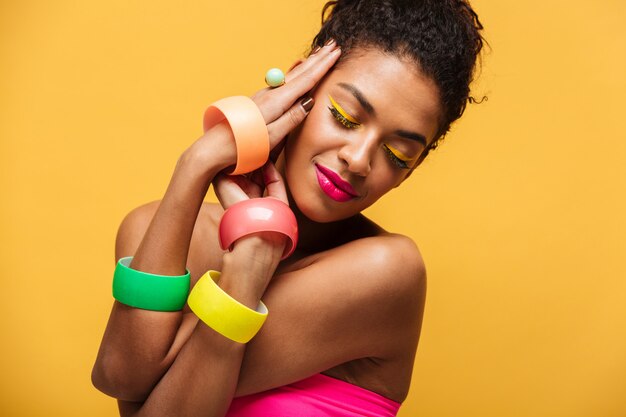 Belle femme afro-américaine élégante avec un maquillage lumineux démontrant des bijoux multicolores se tenant la main au visage isolé, sur jaune