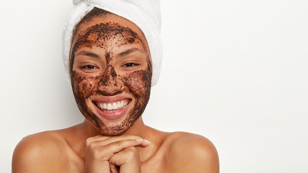 Une belle femme afro-américaine garde les mains jointes, ressent du plaisir et de la joie, se regarde joyeusement dans un miroir, porte une serviette douce enroulée autour de la tête, nettoie le visage avec un masque de beauté.