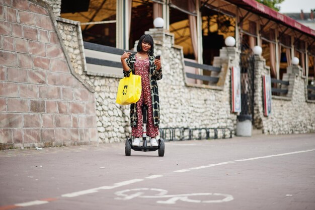 Belle femme afro-américaine utilisant un segway ou un hoverboard et un téléphone portable Fille noire avec symbole de recyclage de sacs écologiques en tissu jaune