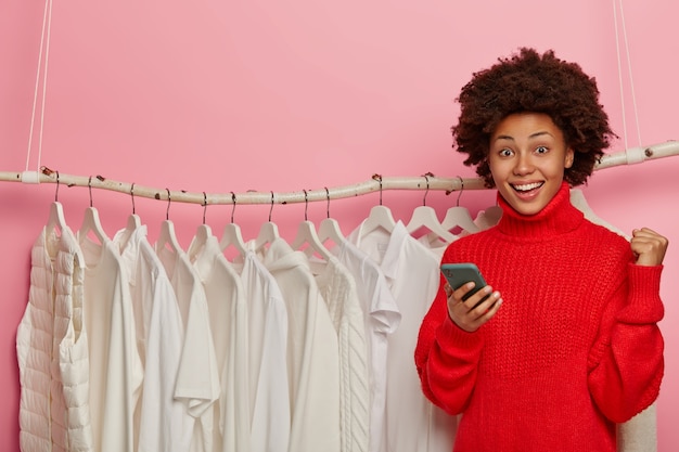 Belle Femme Afro Avec Une Expression Heureuse, Célèbre L'achat Réussi, Se Dresse Contre Des Vêtements Blancs Sur L'espace De Copie De Cintres.