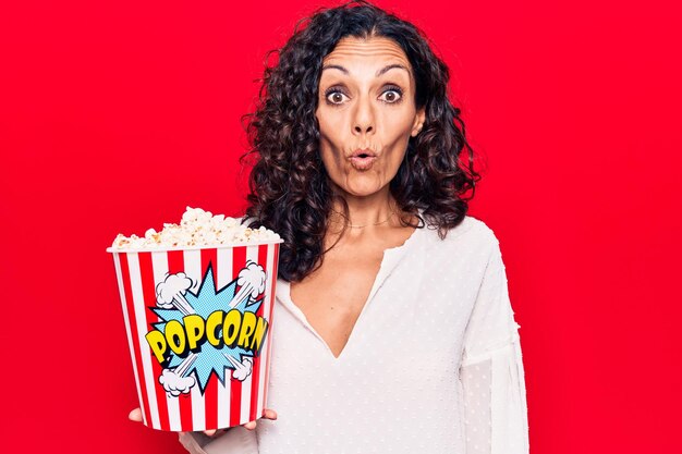 Belle femme d'âge moyen mangeant du pop-corn effrayée et étonnée avec la bouche ouverte pour la surprise, le visage d'incrédulité