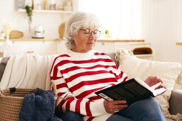 Belle femme âgée aux cheveux gris profitant de sa retraite, assise sur un canapé dans le salon, lisant un roman intéressant. Personnes âgées de race blanche à lunettes rondes de détente à la maison avec livre