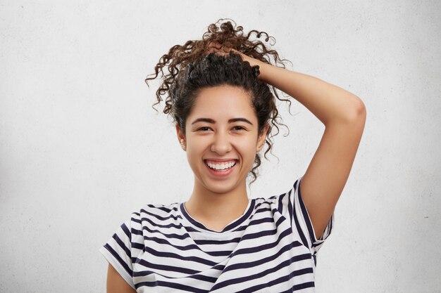 Belle femme agréable à la recherche d'une expression heureuse, ramasse les cheveux bouclés en queue de cheval, s'amuse,