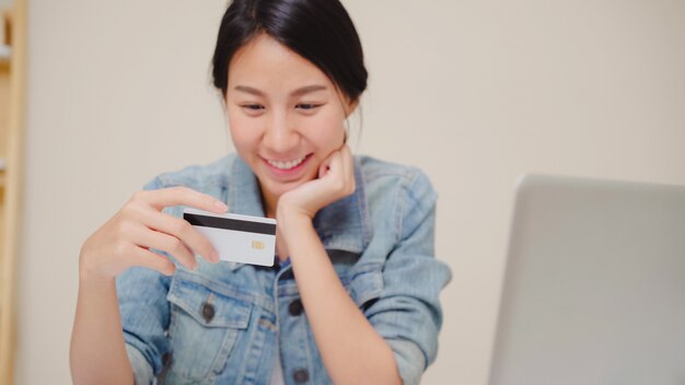 Belle femme asiatique à l&#39;aide d&#39;un ordinateur portable, achats en ligne par carte de crédit tout en tenue décontractée assis sur le bureau dans le salon à la maison.