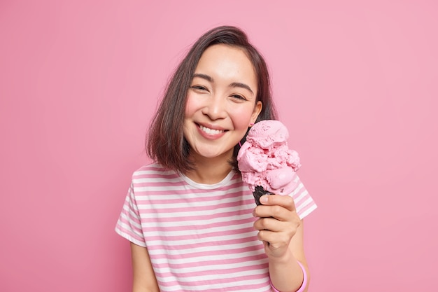 Belle femme asiatique aux cheveux noirs aime manger une délicieuse crème glacée à la fraise pendant les chaudes journées d'été a une humeur optimiste une expression de visage heureuse habillée avec désinvolture pose avec un dessert glacé préféré à l'intérieur