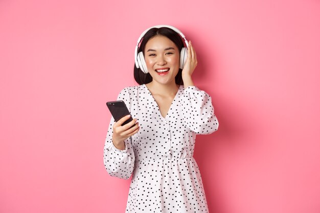 Belle femme asiatique écoutant de la musique dans des écouteurs, utilisant un téléphone portable, souriante heureuse à la caméra, debout sur fond rose