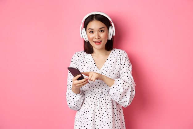 Belle femme asiatique écoutant de la musique dans des écouteurs, utilisant un téléphone portable, souriante heureuse à la caméra, debout sur fond rose