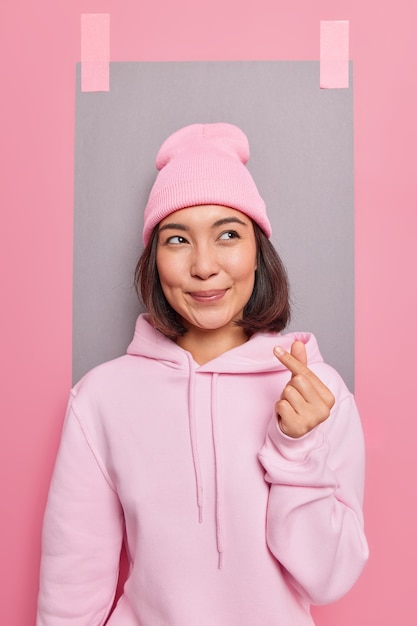 Photo gratuite une belle femme asiatique fait un signe coréen qui montre des sentiments véridiques et l'amour montre un geste de cœur de doigt vêtu d'un sweat à capuche décontracté et pose un chapeau contre le mur du studio avec une feuille de papier plâtrée