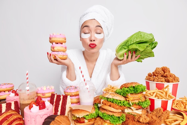 Une belle femme asiatique garde les yeux fermés les lèvres pliées veut vous embrasser tient des beignets et la laitue verte hésite entre les aliments sains et malsains est tentante