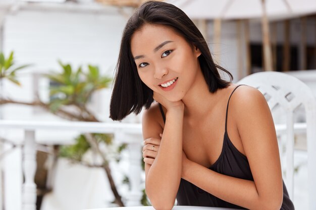 Belle femme asiatique positive avec une expression joyeuse habillée avec désinvolture, assise dans un café en plein air, heureuse d'avoir un bon service recréer avec son amant dans un pays tropical pendant les vacances d'été. Les gens, le style de vie