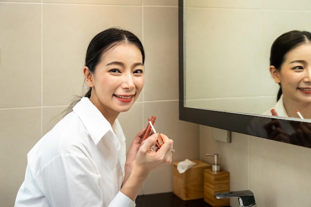 Belle femme asiatique regardant le miroir et appliquant le rouge à lèvres de maquillage de lèvre dans la toilette de salle de bains de salle d'eau