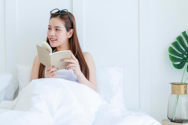 Belle femme asiatique séduisante aime lire un livre sur le lit portrait d'une femme asiatique aux cheveux longs profiter de l'activité du week-end chambre blanche