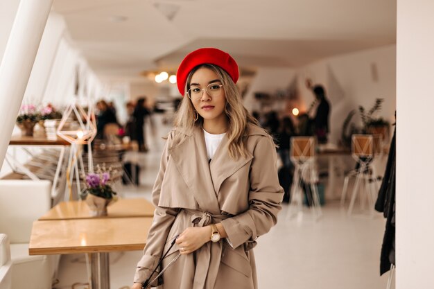 Belle femme asiatique en trench-coat à la mode et béret lumineux tient le sac et regarde à l'avant contre une chambre confortable