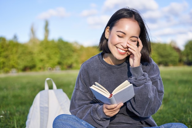 Belle femme assise dans un parc avec un livre souriant fille asiatique lisant et riant se détendre à l'extérieur