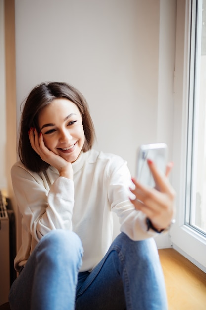 Belle femme assise sur le rebord de la fenêtre en riant et en prenant selfie sur le téléphone