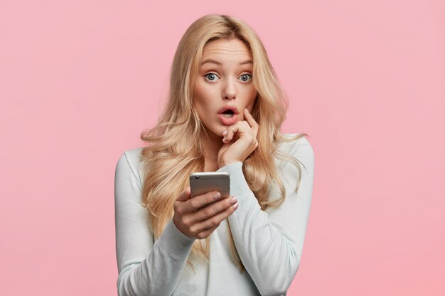 Belle femme aux cheveux blonds se sent surprise en vérifiant sa boîte e-mail sur son téléphone portable, se demandant de gros rabais dans la boutique en ligne, isolée sur un mur rose. Concept d'émotions et d'imprévu