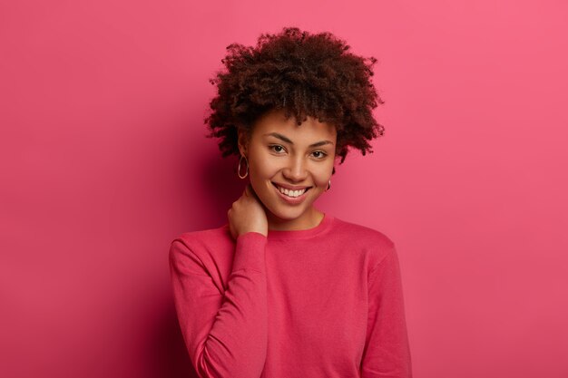 Belle femme aux cheveux bouclés, garde la main sur le cou, sourit doucement, porte un pull décontracté cramoisi, regarde volontiers la caméra