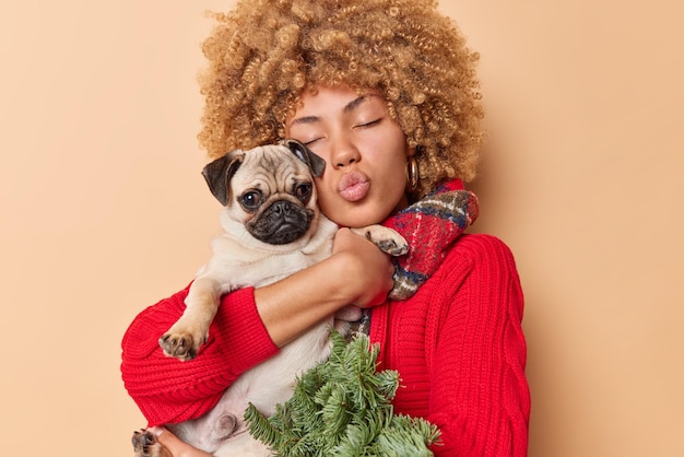 Photo gratuite belle femme aux cheveux bouclés garde les yeux fermés souffle mwah câlins chien de race exprime l'amour vêtu d'un pull rouge tricoté et d'une écharpe autour du cou tient des branches d'épinette verte isolées sur un mur beige