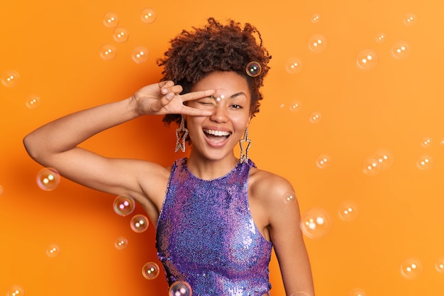 Photo gratuite belle femme aux cheveux bouclés sourit largement vêtue d'une chemise violette vient à la soirée disco fait signe v isolé sur un mur orange volant des bulles de savon reste toujours positif