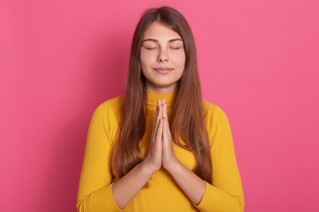 Belle femme aux cheveux longs priant les yeux fermés, gardant la paume ensemble, posant isolée sur un espace rose, portant un pull jaune