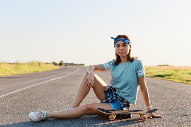 Belle femme aux cheveux noirs portant un t-shirt, un short et des chaussures assis près du skate de surf sur la route goudronnée en plein air, détendez-vous et profitez du surfskate extrême en été.