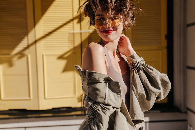 Belle femme aux cheveux ondulés et aux lèvres rouges en veste en denim olive souriant sur les portes jaunes. Dame à lunettes pose sur les portes jaunes