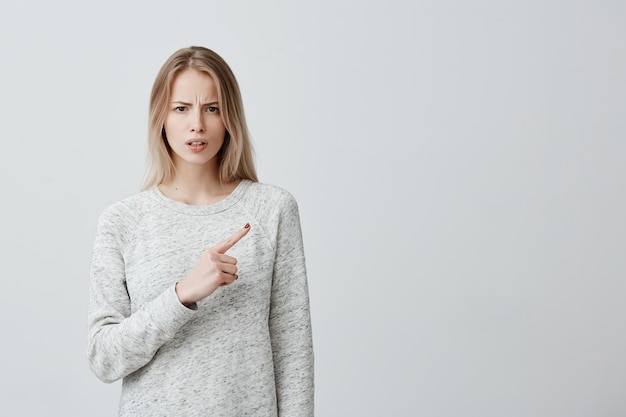 Belle femme aux cheveux teints blonds regardant quelque chose avec dégoût et insatisfaction pointant son index, montrant l'espace de copie