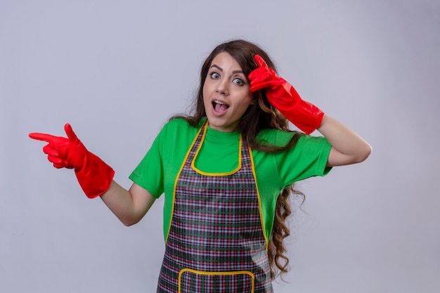 Belle femme aux longs cheveux ondulés portant un tablier et des gants en caoutchouc à la surprise et choqué en pointant avec le doigt sur le côté debout