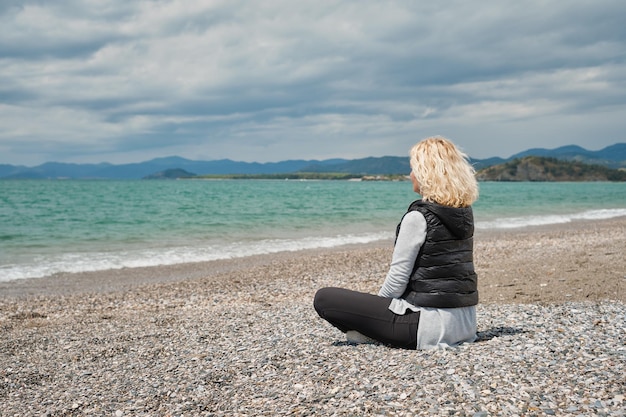 Belle femme blonde adulte en vêtements de sport est assise sur une plage de galets près de la mer Égée azur lumière naturelle du soleil midi Vue de l'arrière sur fond de mer et de vagues vue arrière