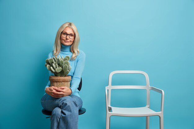 Belle femme blonde d'apparence européenne détient pot de cactus est assis seul près d'une chaise vide étant sur l'isolement à la maison a besoin de communication en direct