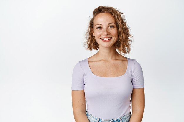 Belle femme blonde avec une courte coupe de cheveux bouclés, l'air sympathique et souriante, debout dans des vêtements décontractés sur blanc.