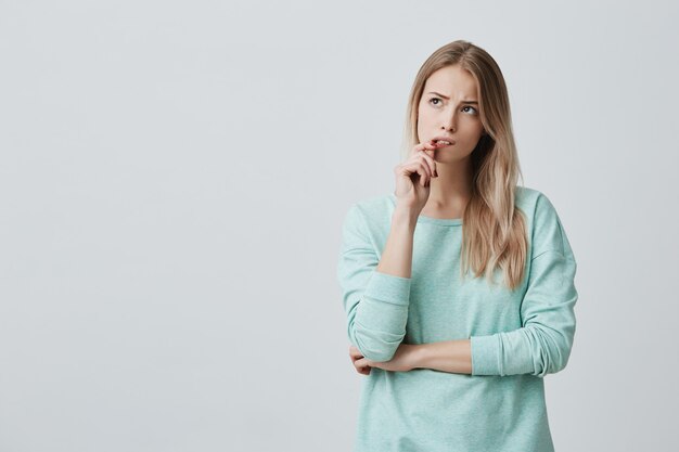 Belle femme blonde avec une expression perplexe, garde le doigt sur les lèvres, regarde de côté avec perplexité