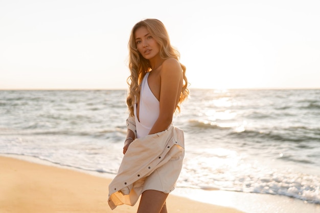 Belle femme blonde en maillot de bain sexy posant sur la plage à la lumière du coucher du soleil.