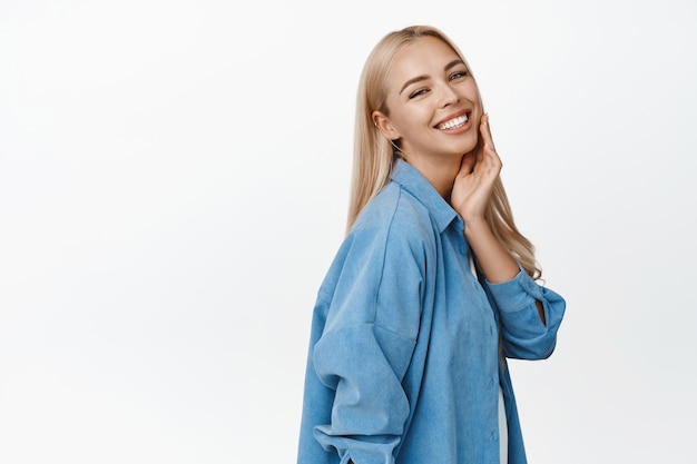 Belle femme blonde avec une peau saine et éclatante souriante heureuse à la caméra touchant son visage profite de la douceur debout sur fond blanc