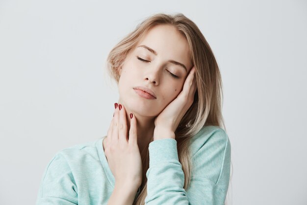 Belle femme blonde pose avec les yeux fermés, touche sa tête avec les mains, a une expression de visage rêveur.