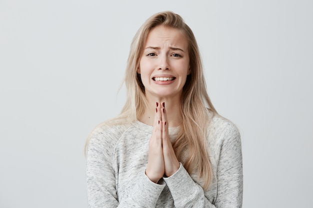 Belle femme blonde serrant les dents avec un regard suppliant tenant les paumes serrées devant elle, demandant pardon, désolé