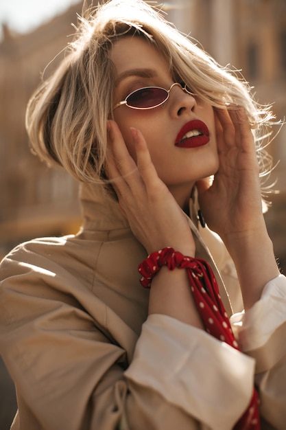 Belle femme blonde sexy confiante en trench-coat beige lunettes de soleil rouges se penche sur la caméra et pose à l'extérieur