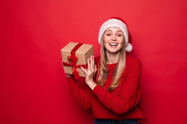 Belle femme en bonnet de noel tenant une boîte-cadeau dans les mains isolées sur un mur rouge